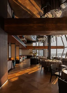 Luxurious multi-level restaurant interior featuring dark wood ceilings, herringbone parquet flooring, round dining tables with neutral linens, and large windows overlooking a waterfront view.