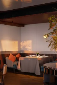 Modern restaurant dining area featuring a round table with grey tablecloth, built-in banquette with orange cushions, walnut wood ceiling beams, and warm ambient lighting casting soft shadows on the wall.