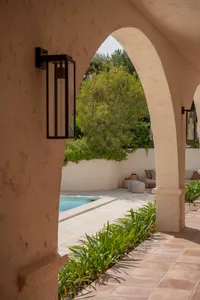 View through a stucco archway into a sunny Mediterranean courtyard featuring a swimming pool, terracotta tile flooring, and abundant green plants.
