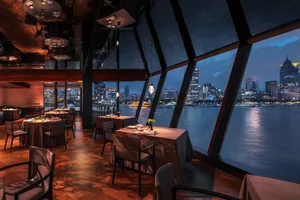 Luxurious modern restaurant interior featuring dark wood beams, herringbone parquet flooring, upholstered dining chairs, and large angled windows revealing a nighttime Shanghai riverside cityscape.