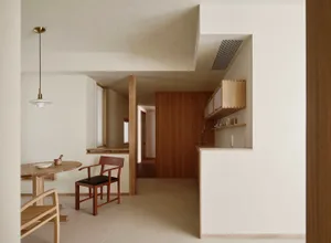 Modern minimalist dining area with round oak table, wooden dining chairs, and a view into a pass-through kitchen featuring light oak cabinetry and white walls.