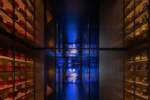 Modern narrow hallway featuring floor-to-ceiling glass and brass shelving filled with illuminated red and amber apothecary jars, dramatic blue ambient lighting, and a large digital firework artwork at the far end.