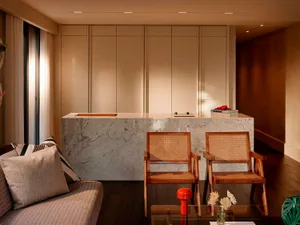 Modern living room featuring a white Carrara marble kitchen island, two wooden rattan armchairs, a beige sofa with striped cushions, and warm recessed lighting.