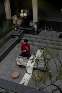 Man in red traditional jacket performing tea ceremony on large natural stone table in a modern stone courtyard at dusk, surrounded by woven cushions and candlelight.