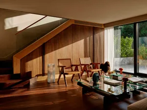 Modern living room featuring warm teak wood paneling, two wooden armchairs with cane backs, a glass coffee table, and dramatic sunlight streaming from a high clerestory window under a staircase.