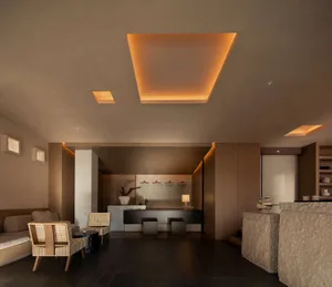 Modern minimalist hotel lobby featuring large travertine stone reception desk, woven rattan armchairs, dark tiled floor, and a dramatic square recessed ceiling with warm orange cove lighting.