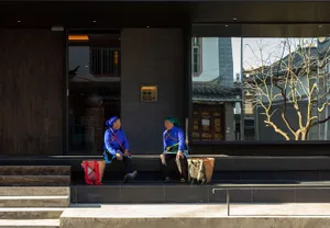 Two elderly women wearing traditional Bai ethnic clothing sit on dark stone steps outside a modern building with large glass windows and dark wood panels, with woven baskets beside them.