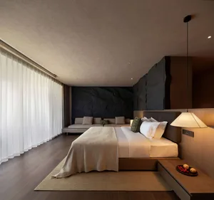 Modern minimalist bedroom with large bed dressed in white and beige linens, dramatic black natural stone feature wall, built-in wooden nightstand, pendant lamp, and sheer white curtains along a full-height window.