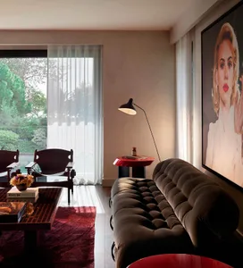 Modern living room with brown tufted velvet sofa, red side table, black floor lamp, wooden lounge chairs, and large Marilyn Monroe portrait on the wall.