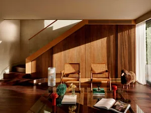 Modern living room featuring a walnut wood wall under a staircase, two rattan armchairs, a glass coffee table with red mushroom stools, and a sheep sculpture beside a window.