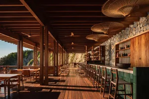 Long open-air wooden veranda with teak flooring and ceiling, featuring a stone wall, green tiled bar counter, woven rattan pendant lamps, and rows of wooden tables and chairs overlooking a dramatic coastal cliff.