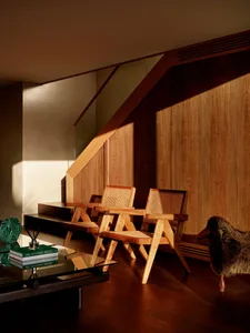 Two wooden armchairs with cane backs placed under a wooden staircase in a warmly lit modern interior with a glass coffee table in the foreground.