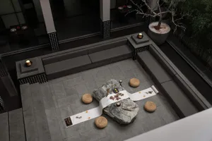 Modern minimalist outdoor courtyard with grey stone pavers, built-in grey benches, large natural rocks, woven straw cushions, and a white embroidered runner set up for a Japanese tea ceremony at night.