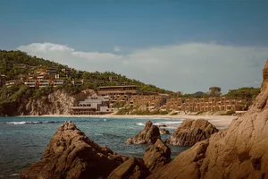 Modern concrete modular resort buildings integrated into a green hillside above a sandy beach and turquoise ocean, viewed from rocky outcrops in the foreground.