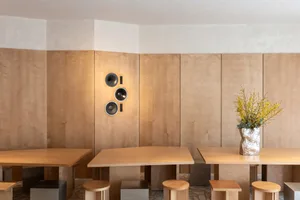 Modern minimalist dining room featuring light oak wood paneling, three sculptural black and silver wall speakers, solid oak rectangular tables, and mixed metal and wood stools, with a vase of yellow forsythia branches.