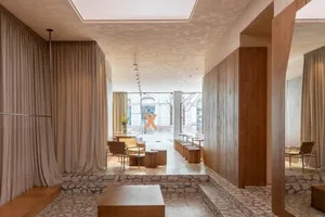 Modern minimalist boutique interior featuring irregular terrazzo flooring and steps, warm oak wood paneling, neutral linen curtains, and contemporary seating areas with wooden tables and leather chairs.