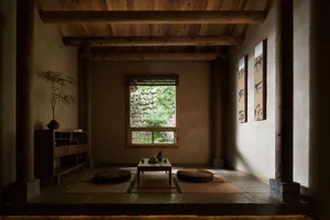 Traditional Japanese tea room interior featuring exposed wooden beams, stone flooring, tatami mats, round zabuton cushions, a low wooden table, and calligraphy scrolls on the wall.