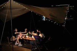 A group of adults and children sitting around a wooden table under a large beige canvas camping tent at night, illuminated by warm string lights, toasting with drinks.
