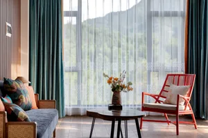 Modern living room featuring a blue sofa with patterned cushions, a bright red rattan armchair, wooden coffee table with flower vase, and large windows overlooking green mountains.