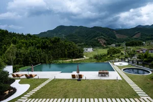Modern infinity pool and concrete terrace with wooden lounge chairs overlooking lush mountains and rural landscape