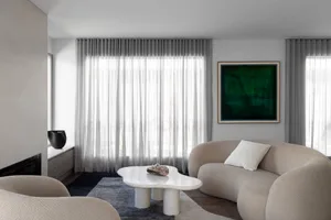 Contemporary living room with beige boucle curved sofa, white irregular marble coffee table, dark blue rug, sheer curtains, and large green abstract painting.
