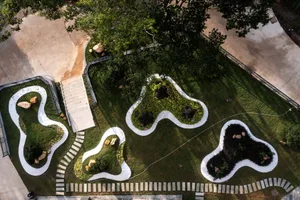 Aerial photograph of a contemporary rooftop garden with organic-shaped planting areas filled with various greenery and rocks, connected by winding white concrete and gravel pathways, surrounded by trees and concrete surfaces.