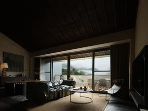 Modern living room featuring a dark wood ceiling, green leather sofa, woven coffee table, chaise lounge, and large glass doors opening to a mountain view with fog.