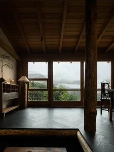 Interior of a wooden mountain cabin featuring large solid wood structural posts and beams, dark herringbone wood flooring, minimalist wooden furniture, and expansive windows overlooking misty forested mountains.