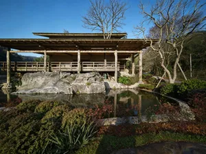 Traditional Japanese wooden pavilion built over a reflective koi pond surrounded by large natural rocks, manicured shrubs, and bare winter trees under a clear blue sky.