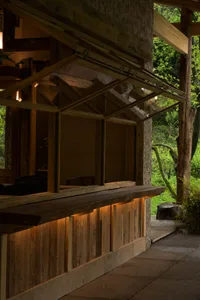 Japanese-style outdoor wooden bar counter with uplighting, set against a dense green forest backdrop under a timber pavilion structure.