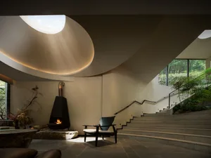 Modern minimalist living space featuring a sculptural black fireplace on a stone platform, a dark wood armchair, large stone coffee table, and a sweeping curved ceiling with a prominent circular skylight that casts beautiful light rays. A wide stone staircase ascends to the right with forest views beyond.