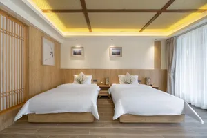 Modern twin hotel room with two white beds, light oak wood walls and flooring, woven bamboo ceiling with warm cove lighting, and framed nature artwork.