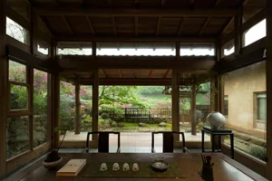 Interior view of a traditional Japanese tea room with dark wooden beams, large glass windows, and a direct view of a lush garden with pink azaleas and a wooden veranda.
