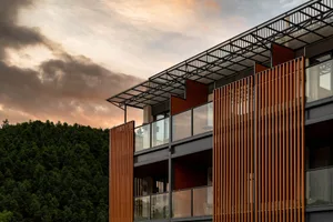 Modern multi-story residential building with vertical cedar wood cladding, large glass windows, and metal railings against a dense green forest at sunset.