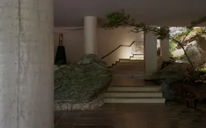 Modern brutalist entrance hall featuring large natural stone boulders, concrete pillars, a curving staircase, and integrated indoor trees under soft ambient lighting.