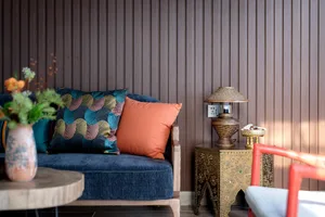 Blue velvet sofa with colorful patterned and solid orange cushions, placed next to an ornate brass side table with a woven lamp against a dark wood slatted wall.