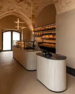 Modern bakery interior featuring a curved fluted oak service counter with black marble top, glass pastry display case, and historic stone vaulted ceiling with modern linear pendant light.