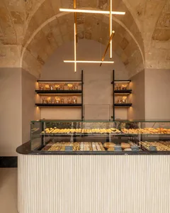 Modern bakery counter with glass pastry display filled with croissants and pastries, set against historic stone vaulted ceiling and illuminated floating shelves.