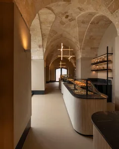 Interior view of a contemporary bakery housed within a historic stone-vaulted space featuring a curved wooden pastry display counter and modern geometric pendant lights.