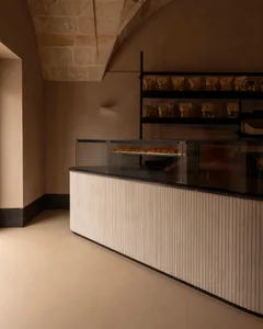 Modern curved bakery counter with vertical fluted wood paneling, glass pastry display case, and black metal shelving against a backdrop of original stone vaulted ceiling.