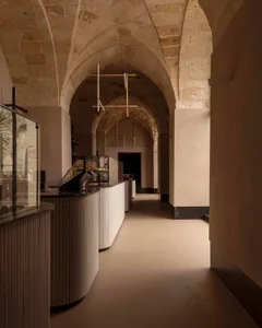Long view down a narrow bakery counter with fluted walnut paneling and black marble top, set beneath dramatic historic limestone vaulted ceilings with modern geometric pendant lights.