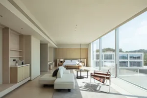 Modern minimalist bedroom with light oak cabinetry, white sectional sofa, brown leather sling chair, and floor-to-ceiling glass windows overlooking trees.