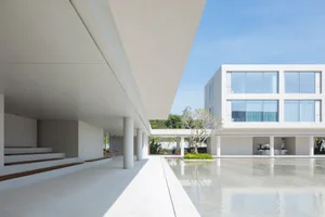 Modern minimalist concrete building with large reflecting pool in a sunlit courtyard featuring white pillars and a small tree.