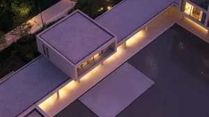 Aerial night view of a modern minimalist concrete villa with flat gravel roof, dramatic linear LED lighting along the eaves, and a large reflecting pool.