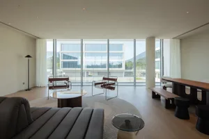 Modern minimalist living room featuring brown leather and chrome lounge chairs, walnut coffee tables, a curved rug, and large floor-to-ceiling windows overlooking mountains and contemporary buildings.