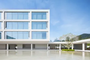 Modern minimalist concrete building with large glass windows, elevated on pillars, overlooking a calm reflecting pool with a single tree and mountain view in the background.