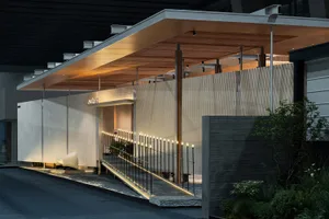 Night view of a modern minimalist commercial building entrance with concrete walls, vertical wood paneling, a cantilevered warm wood canopy, illuminated ramp railing, and a white sculptural chair.