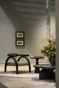 Modern minimalist entryway featuring a black arched accent chair with textured seat, round black side table, large floral arrangement in a blue ceramic vase, and two framed artworks on a grey concrete wall.