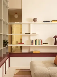 Modern minimalist living room featuring white floating shelves, burgundy lacquered cabinetry, a beige modular sofa, and warm lighting with a striped ceramic vase and colorful books.