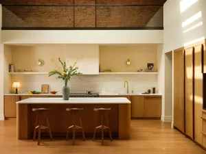 Modern open-plan kitchen with a large central oak island, white marble countertops, three wooden stools, and exposed brick loft wall above.
